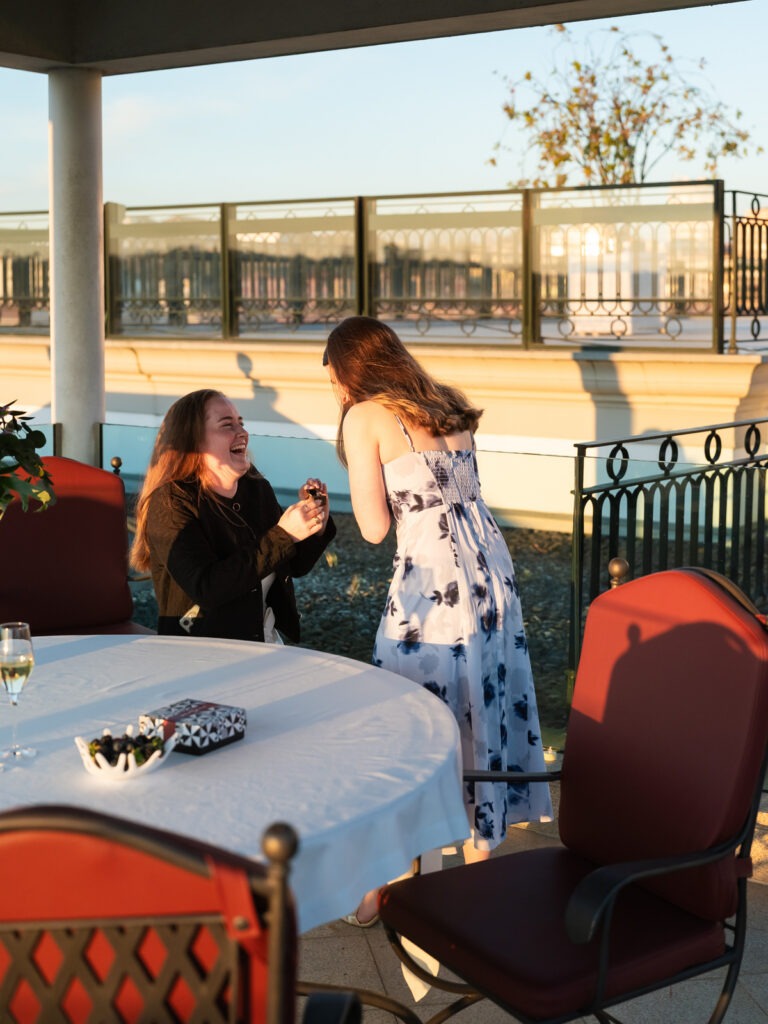 Pedido de casamento ao pôr do sol, uma noiva ajoelha-se e pede em casamento, enquanto a outra, de costas leva as mãos à boca surpreendida. 