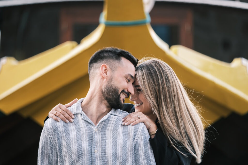 Casal em sessão pré-casamento no Cais de Gaia; momento cúmplice em exterior, com barco desfocado ao fundo e luz natural suave junto ao Douro.